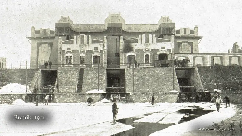 Black-and-white 1913 photograph of the Braník ice house above its stone embankment, with the film's lower third Braník, 1911